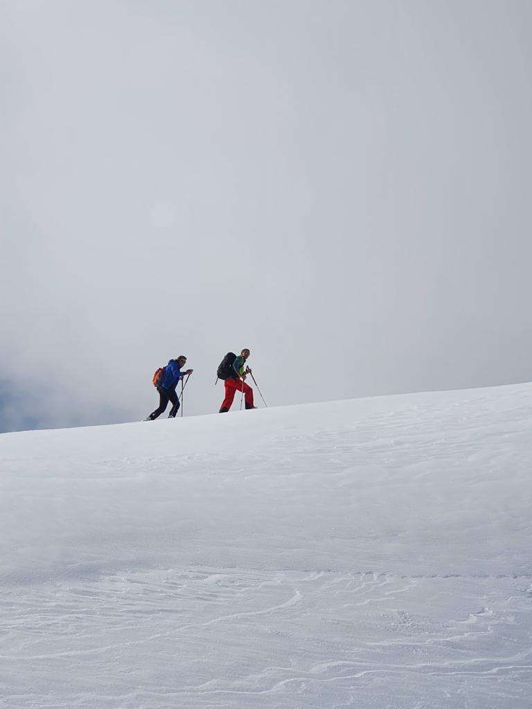 Deux randonneurs équipés de sacs à dos et de bâtons de marche montent une pente enneigée sous un ciel gris.